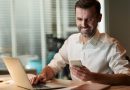 Homem sorrindo enquanto usa celular e computador ao mesmo tempo