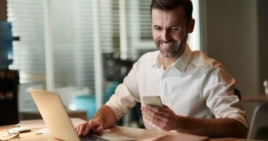 Homem sorrindo enquanto usa celular e computador ao mesmo tempo